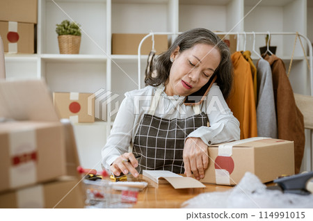 A mature Asian female tailor is taking notes on a notepad and receiving orders from the call. A mature Asian female tailor is taking notes on a notepad and receiving orders from the call. 114991015