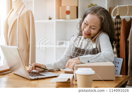 A cheerful, busy mature Asian female tailor or online seller is working in her atelier studio. 114991026