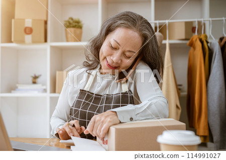A cheerful, busy mature Asian female tailor or online seller is working in her atelier studio. 114991027