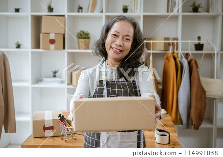 An attractive mature Asian female online seller is holding a cardboard box and smiling at the camera An attractive mature Asian female online seller is holding a cardboard box and smiling at the camera 114991038
