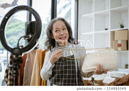 A mature Asian female online seller is live streaming from home, selling her products online. A mature Asian female online seller is live streaming from home, selling her products online. 114991049