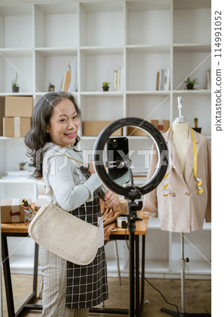 A mature Asian female online seller is live streaming from home, selling her products online. 114991052