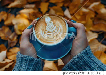 Female hand in sweater holding coffee cup with beautiful latte art outdoors. Autumn aesthetic concept. Cappuccino coffee blue mug, yellow fall leaves background. Cozy autumnal composition. Hot drink 114991316