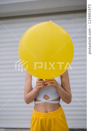 Cheerful young woman wearing yellow pants hiding behind yellow balloon Cheerful young woman wearing yellow pants hiding behind yellow balloon 114991973