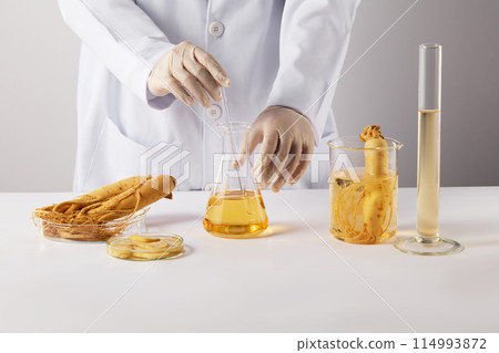 Front view photo on white texture, a researcher holds a glass stick stirring yellow solution in erlenmeyer flask placed beside a beaker, measuring cylinder and petri dishes of ginseng on white table 114993872