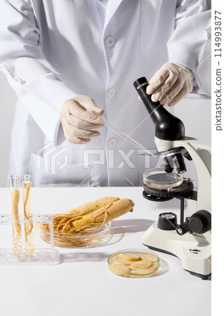 The researcher holds a microscope in one hand and a dropper in the other hand, preparing to add more solution to the petri dish placed on the microscope, placed on white table next to other lab items The researcher holds a microscope in one hand and a dropper in the other hand, preparing to add more solution to the petri dish placed on the microscope, placed on white table next to other lab items 114993877