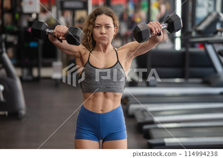Muscular Caucasian forty year old woman doing exercises with dumbbells in the gym. Muscular Caucasian forty year old woman doing exercises with dumbbells in the gym. 114994238