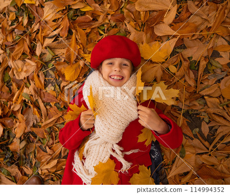 An overhead view of a caucasian girl in a red coat and beret lies on yellow foliage. Walk in the park in autumn.  114994352
