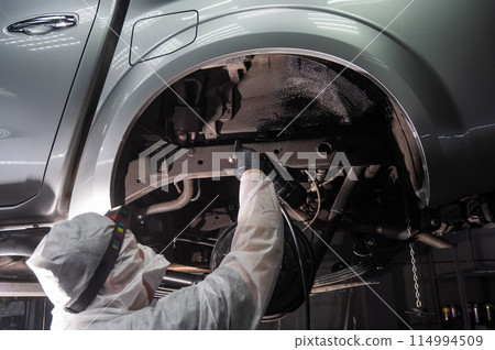An auto mechanic applies anti-corrosion mastic to the underbody of a car. An auto mechanic applies anti-corrosion mastic to the underbody of a car. 114994509