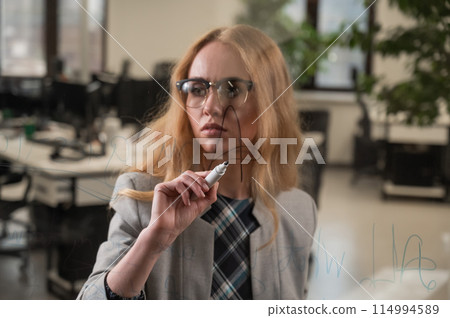 Caucasian woman with glasses writes text in English on a glass wall.  114994589