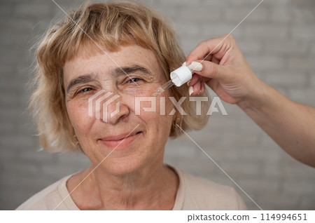 Close-up portrait of an old woman applying hyaluronic acid serum with a pipette. Anti-aging face care.  114994651