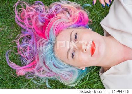 Top view of Caucasian woman with multi-colored hair lying on green grass. Top view of Caucasian woman with multi-colored hair lying on green grass. 114994716