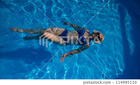 An elderly woman in sunglasses swims on her back in the pool. Vacation in retirement. An elderly woman in sunglasses swims on her back in the pool. Vacation in retirement. 114995110