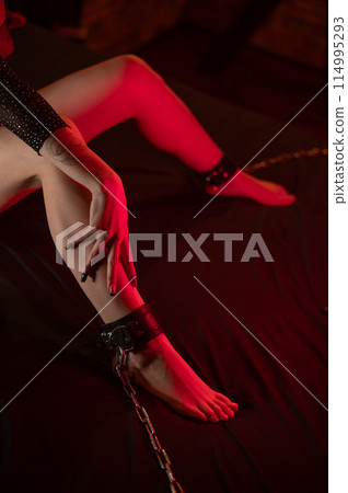 A faceless woman sits on a bed with her ankles handcuffed. BDSM concept. Vertical photo.  114995293