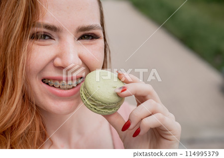 Young red-haired woman with braces eating macaron cake. Young red-haired woman with braces eating macaron cake. 114995379