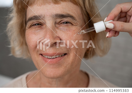 Close-up portrait of an old woman applying hyaluronic acid serum with a pipette. Anti-aging face care. Close-up portrait of an old woman applying hyaluronic acid serum with a pipette. Anti-aging face care. 114995383