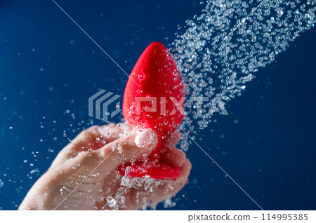 Woman washing red anal plug under shower on blue background. Woman washing red anal plug under shower on blue background. 114995385