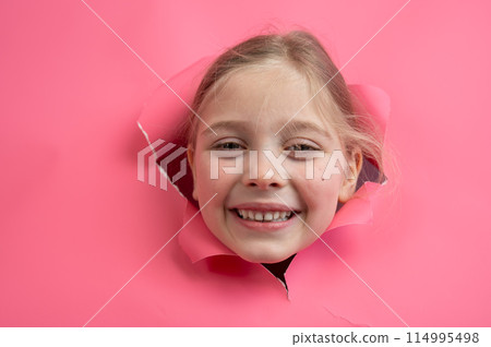 Cute Caucasian girl peeks out of a hole in a paper pink background. 114995498