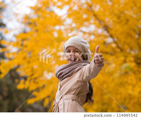 Happy caucasian girl in a beige coat and beret walks in the park in autumn. Schoolgirl showing thumbs up. Happy caucasian girl in a beige coat and beret walks in the park in autumn. Schoolgirl showing thumbs up. 114995542