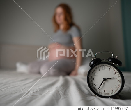 Caucasian pregnant woman sits on the bed and suffers from insomnia. Alarm clock in the foreground. Caucasian pregnant woman sits on the bed and suffers from insomnia. Alarm clock in the foreground. 114995597