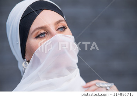 Portrait of a young blue-eyed woman in a hijab against a gray brick wall. Muslim woman with white scarf covering her face.  114995713
