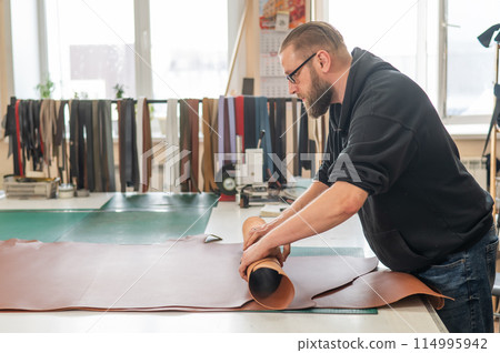Leatherworker unrolls rolls of leather in workshop. Leatherworker unrolls rolls of leather in workshop. 114995942