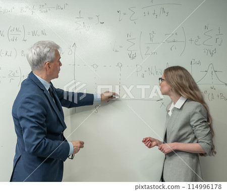 An elderly professor explains a subject to a student at a white board. An elderly professor explains a subject to a student at a white board. 114996178