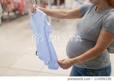 Caucasian pregnant woman chooses baby clothes in a store. Faceless expectant mother in the 3rd trimester.  114996190