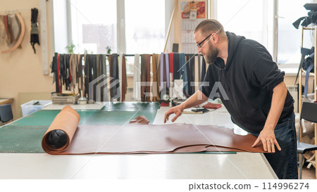 Leatherworker unrolls rolls of leather in workshop. Leatherworker unrolls rolls of leather in workshop. 114996274