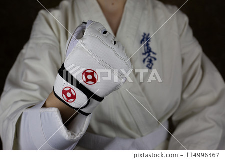Girl athlete in Kyokushinkai karate demonstrates clenched fist in special protective glove for martial arts. Athlete in karate kimono. Inscription in Japanese: Kyokushinkai 114996367