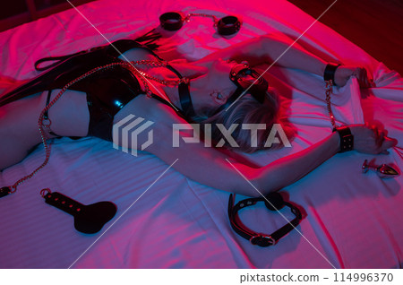 Sexy blonde woman in a leather mask and handcuffs and a collar lies in the bedroom in a red-blue neon light. Role-playing games. Sexy blonde woman in a leather mask and handcuffs and a collar lies in the bedroom in a red-blue neon light. Role-playing games. 114996370