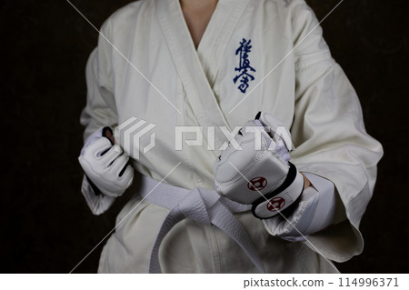 Young woman in Kyokushin karate kimono in fighting stance with lowered clenched fists in special protective gloves in front of her opponent. Inscription in Japanese: Kyokushinkai Young woman in Kyokushin karate kimono in fighting stance with lowered clenched fists in special protective gloves in front of her opponent. Inscription in Japanese: Kyokushinkai 114996371
