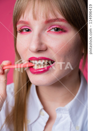 Portrait of a young woman with braces and bright makeup chewing gum on a pink background. Vertical photo. Portrait of a young woman with braces and bright makeup chewing gum on a pink background. Vertical photo. 114996419