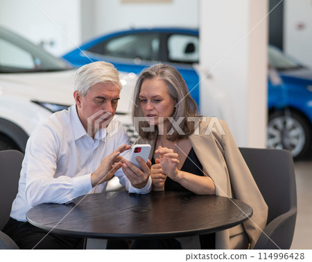 A mature couple is sitting in a car dealership and looking at a smartphone. A mature couple is sitting in a car dealership and looking at a smartphone. 114996428