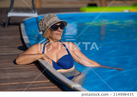 An elderly woman in a hat and sunglasses swims in the pool. Retirement leave.  114996435
