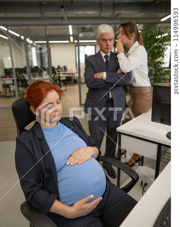Elderly Caucasian man and woman gossiping behind their sleeping pregnant colleague in the office. Vertical photo.  114996593