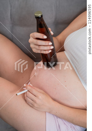 Faceless pregnant woman drinks beer while sitting on the sofa.  114996595