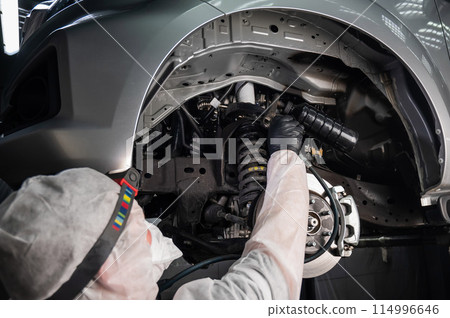 An auto mechanic applies anti-corrosion mastic to the underbody of a car. 114996646