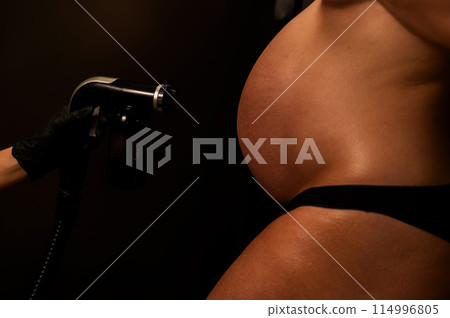 Close-up of the belly of a pregnant woman undergoing an instant tanning procedure. The master applies bronzer with a spray. Close-up of the belly of a pregnant woman undergoing an instant tanning procedure. The master applies bronzer with a spray. 114996805