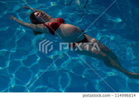 Top view of pregnant woman floating in pool in red bikini.  114996814