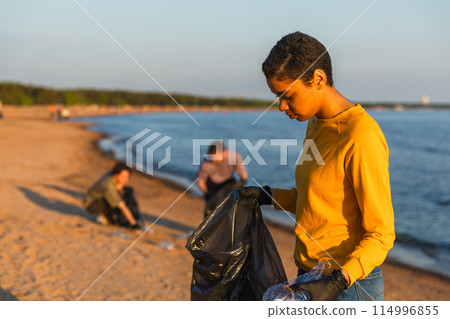Earth day. Volunteers activists team collects garbage cleaning of beach coastal zone. Woman puts plastic bottle trash in garbage bag on ocean shore. Environmental conservation coastal zone cleaning Earth day. Volunteers activists team collects garbage cleaning of beach coastal zone. Woman puts plastic bottle trash in garbage bag on ocean shore. Environmental conservation coastal zone cleaning 114996855