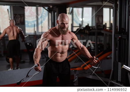 Bald Caucasian bodybuilder training chest using cable crossover in gym.  114996932