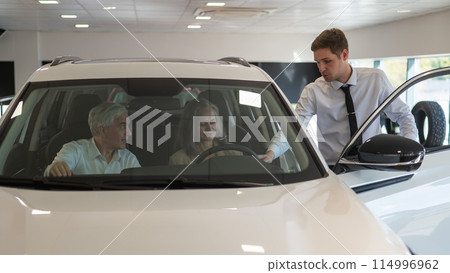 A gray-haired Caucasian woman is sitting behind the wheel of a car. Car dealership salesman consulting mature married couple. 114996962
