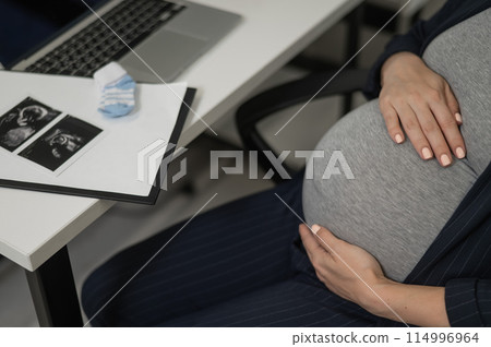 A pregnant woman works on a laptop in the office and looks at a photo from an ultrasound scan of the fetus. Belly close-up. 114996964