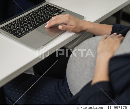 A pregnant woman works on a laptop in the office. Close-up of the tummy. A pregnant woman works on a laptop in the office. Close-up of the tummy. 114997176