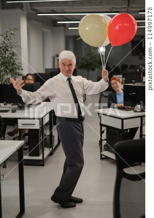 Portrait of a cheerful mature business man holding balloons in the office. Vertical photo.  114997178