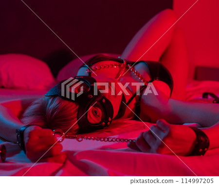 Sexy blonde woman in a leather mask and handcuffs and a collar lies in the bedroom in a red-blue neon light. Role-playing games. Sexy blonde woman in a leather mask and handcuffs and a collar lies in the bedroom in a red-blue neon light. Role-playing games. 114997205