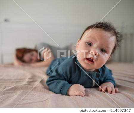 A three-month-old boy lies on his stomach on the bed and his mother sleeps behind him. Postpartum depression. 114997247