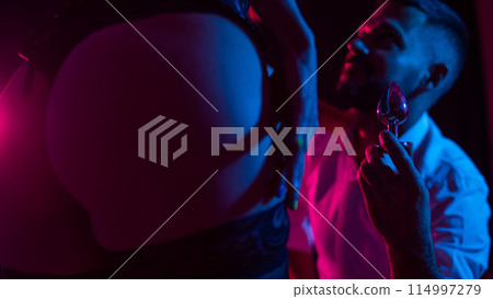 Bearded caucasian man holding an anal plug. Close-up of female buttocks in underwear. Sexual preferences of the couple. 114997279