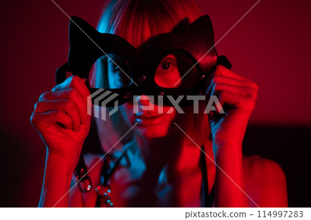 Blonde woman putting on leather mask in bedroom in red blue neon light. Blonde woman putting on leather mask in bedroom in red blue neon light. 114997283
